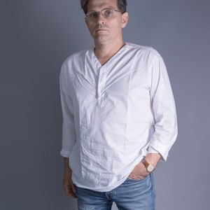 May include: A man wearing a white long-sleeved shirt with a button-up collar and blue jeans. He is wearing glasses and a watch on his left wrist.
