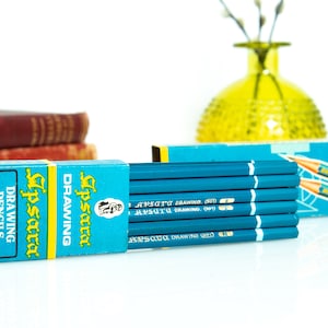 May include: A set of blue drawing pencils with white stripes and text, packaged in a blue and yellow box. The box and pencils have the text "Apsara Drawing Pencils". A yellow vase and books are in the background.