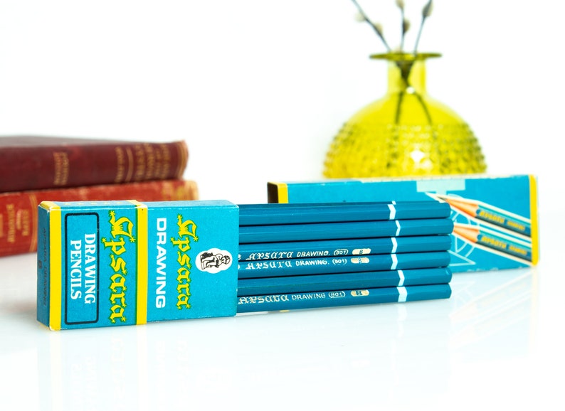 May include: A set of blue drawing pencils with white stripes and text, packaged in a blue and yellow box. The box and pencils have the text "Apsara Drawing Pencils". A yellow vase and books are in the background.