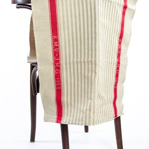 Könnte beinhalten: Ein gestreiftes beige-rotes Geschirrtuch mit dem aufgedruckten Text "K.M.V.-J.M.G. 1953". Das Tuch ist über einen Holzstuhl gelegt.