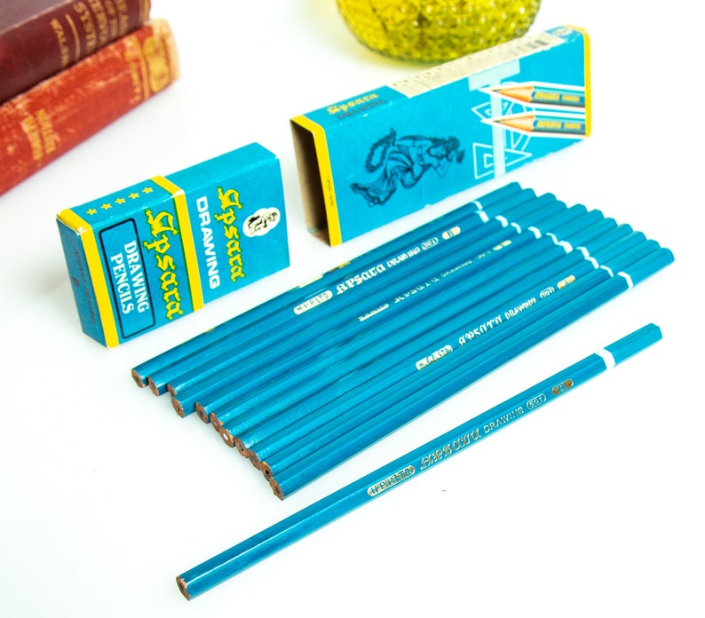 May include: A collection of blue drawing pencils with white tips and the text "Apsara Drawing 601". Two blue boxes with the text "Lipsara Drawing Pencils" are also present. The pencils are arranged on a white surface.