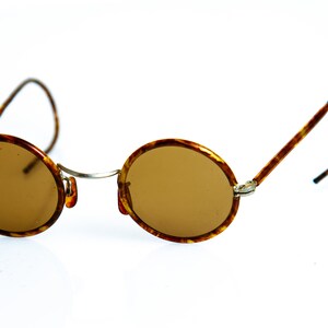 May include: A pair of round, brown tortoise shell sunglasses with brown lenses. The sunglasses have a thin metal frame and are sitting on a white surface.