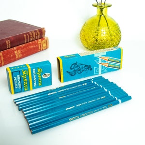 May include: A collection of blue drawing pencils with matching boxes. The boxes are teal with yellow accents and feature the text "Apsara Drawing Pencils". Two vintage books and a green glass vase are in the background.