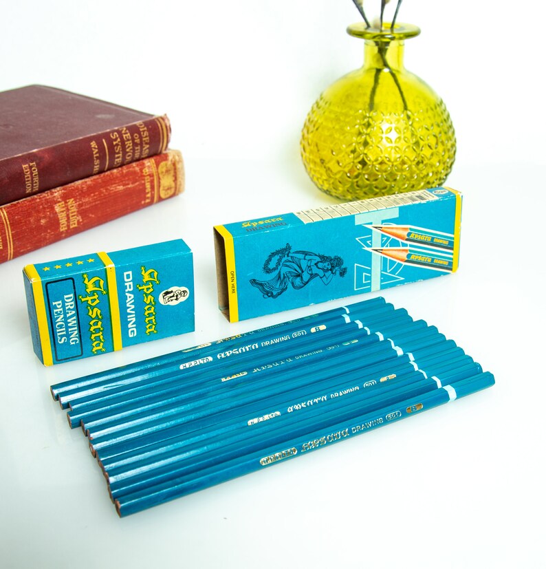 May include: A collection of blue drawing pencils with matching boxes. The boxes are teal with yellow accents and feature the text "Apsara Drawing Pencils". Two vintage books and a green glass vase are in the background.
