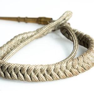 May include: A braided cord with a gold-colored metal tip. The cord is a light beige color and is braided in a complex pattern.
