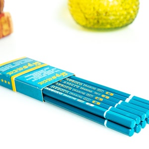 May include: A partially open box of teal drawing pencils with the text "Apsara Drawing" visible. The pencils are teal with white and gold accents. The box is teal and yellow with text and star details.