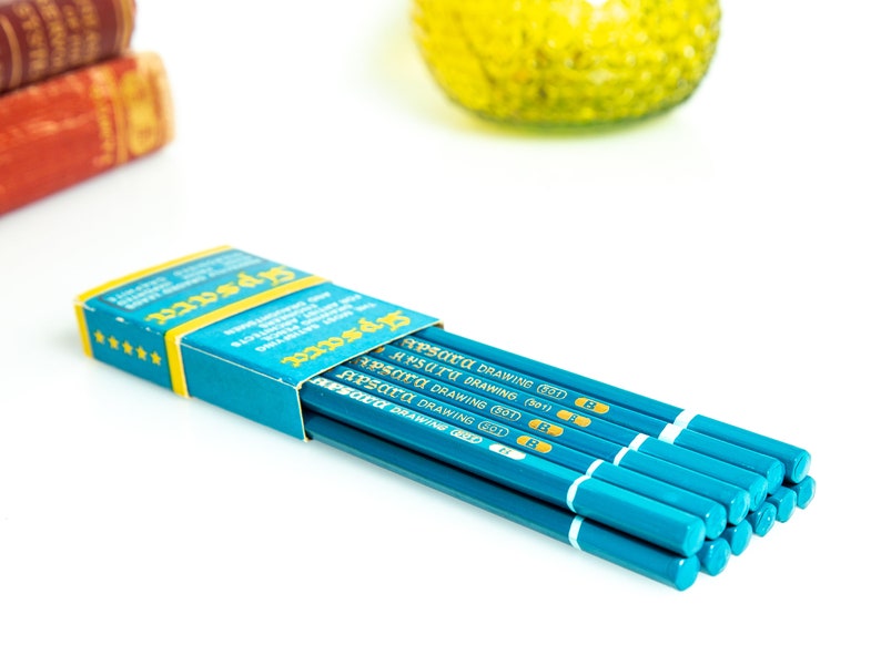 May include: A partially open box of teal drawing pencils with the text "Apsara Drawing" visible. The pencils are teal with white and gold accents. The box is teal and yellow with text and star details.