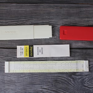 May include: A vintage slide rule set, including a white case with "ARISTO" printed on it, a red case, a manual, and a slide rule. The slide rule has yellowed scales and white markings. The set is on a dark wood surface.