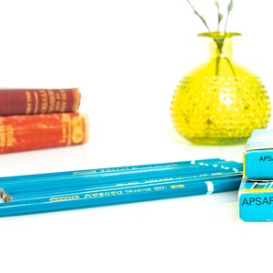May include: A still life arrangement featuring blue pencils, two boxes of Apsara Drawing B pencils, and two vintage books with red covers. A yellow glass vase with a textured surface is also present.