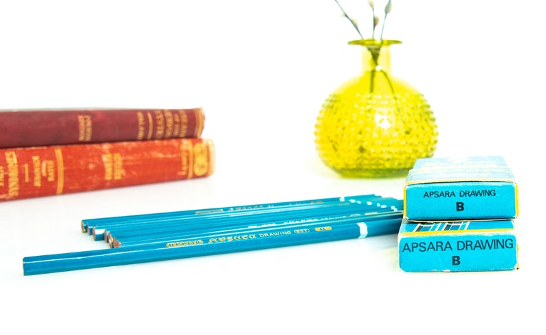May include: A still life arrangement featuring blue pencils, two boxes of Apsara Drawing B pencils, and two vintage books with red covers. A yellow glass vase with a textured surface is also present.