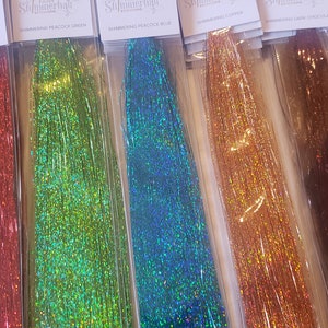 May include: A selection of shimmering hair tinsel in various colors, including gold, red, green, blue, copper, dark chocolate, and pink. Each package is labeled with the color name and is packaged in a clear plastic sleeve. The tinsel strands are long and glittery.