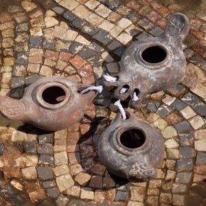May include: Three terracotta oil lamps with white wicks are positioned on a mosaic tiled floor. The lamps are of different shapes and sizes, with one featuring a handle.