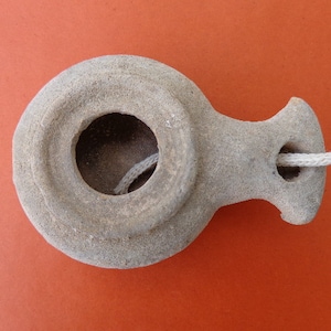 Puede incluir: Una lámpara de aceite de cerámica gris con un depósito redondo y un asa. La lámpara tiene un pequeño agujero en el centro del depósito para una mecha.