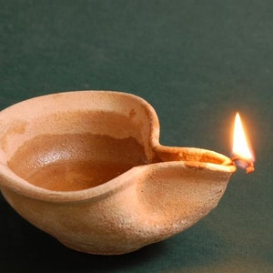Puede incluir: Una l&aacute;mpara de aceite de terracota con una peque&ntilde;a llama que arde en su interior. La l&aacute;mpara est&aacute; hecha de arcilla rugosa y texturizada y tiene un pico en el lateral.