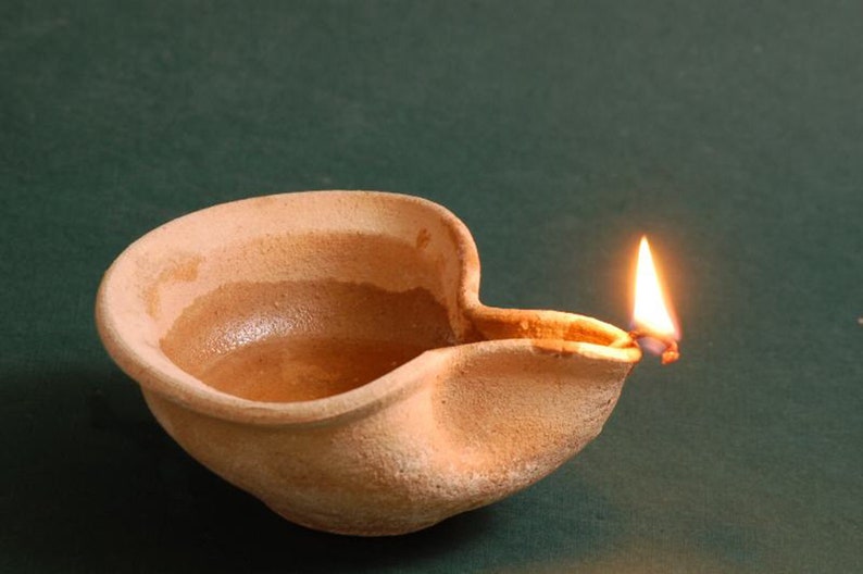 Puede incluir: Una l&aacute;mpara de aceite de terracota con una peque&ntilde;a llama que arde en su interior. La l&aacute;mpara est&aacute; hecha de arcilla rugosa y texturizada y tiene un pico en el lateral.