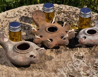 May include: Three small, brown clay oil lamps with a small glass bottle of oil next to each. The lamps are all different shapes and sizes, and the bottles are all the same size. The lamps are sitting on a rough, brown surface.