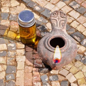 Può includere: Una piccola bottiglia di vetro piena di un liquido dorato si trova accanto a una lampada ad olio in terracotta con una fiamma che brucia all'interno. La lampada ha un design a forma di croce sul manico. Entrambi gli oggetti sono su un pavimento a mosaico.
