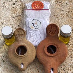 May include: Two terracotta oil lamps with wicks and a small bottle of olive oil. The lamps are brown and beige and have a cross design on the top. The bottle is clear glass with a silver lid. The image also includes a bag of white cotton wicks with a sticker that says "Eco friendly candles Light Lamps".