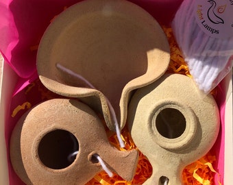May include: Three terracotta oil lamps in a pink gift box with yellow shredded paper. The lamps are in various shapes and sizes. The box also includes a package of white cotton wicks labeled "Light Lamps Eco friendly".