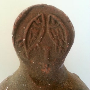 May include: A brown, textured, circular clay object featuring an embossed design of two fish facing each other, positioned above a small cross-like symbol. The surface has a rough, earthy appearance with visible specks of lighter material.