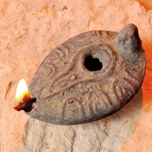 May include: An antique, oval-shaped oil lamp made of weathered brown clay. The lamp features a central hole and is adorned with etched patterns. A lit wick emits a bright yellow flame, casting a warm glow. The lamp rests on a textured, light brown surface.