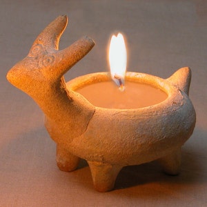 Puede incluir: Un portavelas de cer&aacute;mica con forma de ciervo con una vela encendida en su interior. El portavelas es de color marr&oacute;n claro y tiene una textura rugosa.