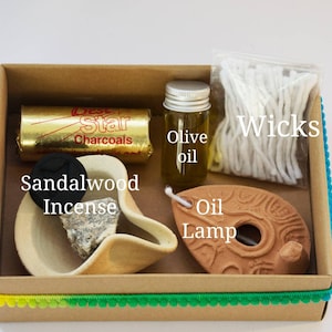 May include: A brown cardboard box with a colorful trim contains a set of items for burning incense and oil. The items include a small bottle of olive oil, a clay oil lamp, a pack of wicks, a block of sandalwood incense, and a pack of charcoal briquettes. The box is labeled "Best Star Charcoals".