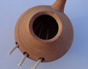 Puede incluir: Una lámpara de aceite de terracota con cuerpo redondo y un pico. La lámpara tiene cuatro agujeros alrededor de la base, con mechas de algodón blanco enhebradas a través de ellos.