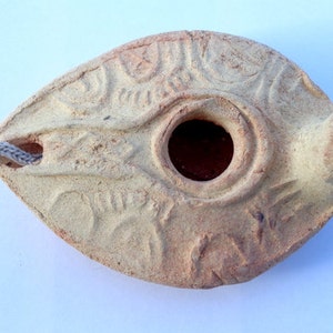 May include: A small, light brown clay oil lamp with intricate carvings. The lamp has a round hole in the center for the wick and a spout on the side for pouring oil.