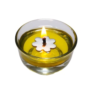May include: A glass candle holder containing yellow liquid and a white flower-shaped wick. The wick is lit and the flame is visible.