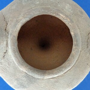 May include: A terracotta oil lamp with a circular opening. The lamp is made of a rough, textured clay and has a light brown color.