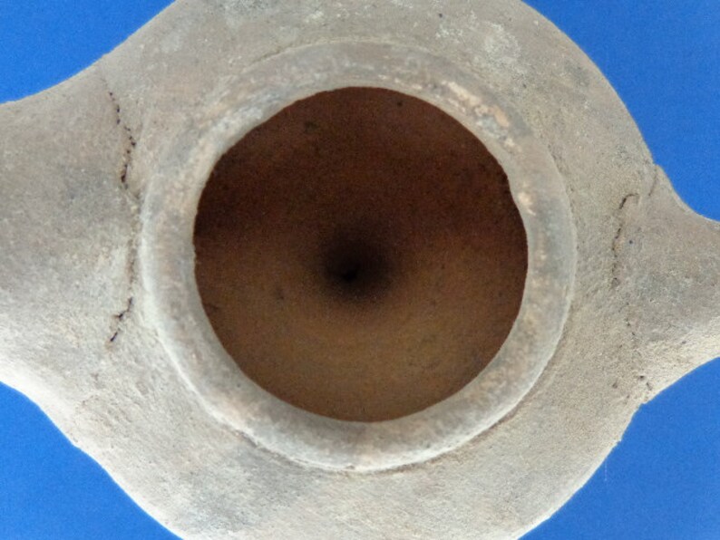 May include: A terracotta oil lamp with a circular opening. The lamp is made of a rough, textured clay and has a light brown color.