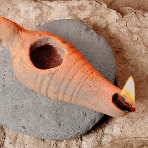 Könnte beinhalten: Eine terracotta-Öllampe mit einer brennenden Flamme, die auf einem grauen Stein ruht. Die Lampe hat eine runde Öffnung und einen Ausguss an der Seite.