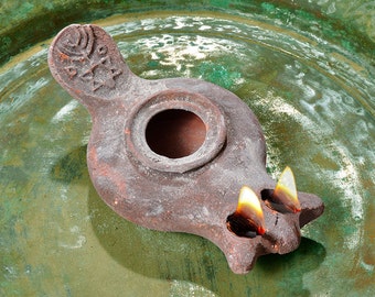 May include: A terracotta oil lamp with two wicks burning, sitting on a green metal surface. The lamp has a round opening in the center and a handle with a design on it.