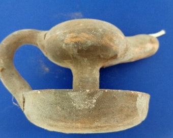 Puede incluir: Una lámpara de aceite de terracota con asa y boquilla. La lámpara está en mal estado, con un color marrón claro.
