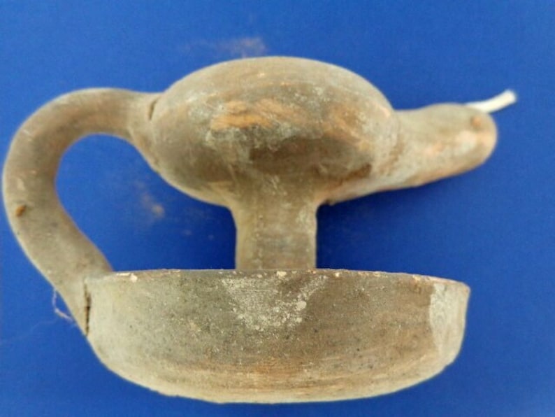May include: A terracotta oil lamp with a handle and a spout. The lamp is in a worn condition, with a light brown color.
