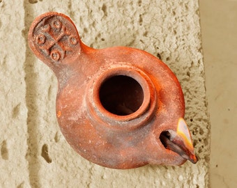 Puede incluir: Una lámpara de aceite de terracota con cuerpo circular y una abertura central. El asa presenta un diseño de cruz en relieve. Una pequeña llama es visible en la boquilla que se extiende desde el lateral del recipiente de arcilla rojiza.