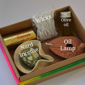 May include: A brown cardboard box with a colorful trim contains a set of items for burning incense. The box includes a small clay oil lamp, a small bowl of Nard incense, a package of wicks, a small bottle of olive oil, and a package of charcoals. The box is labeled "Charcoals" in Arabic.