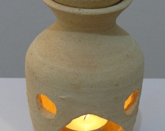 Puede incluir: Un difusor de aceite de cerámica beige con una parte superior redonda y una vela pequeña en el interior. El difusor tiene dos agujeros en los lados y una abertura más grande en la parte delantera para la vela.