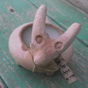 Puede incluir: Una maceta de terracota con forma de animal estilizado con una vela blanca en su interior. La maceta tiene una peque&ntilde;a etiqueta adjunta con texto en hebreo.
