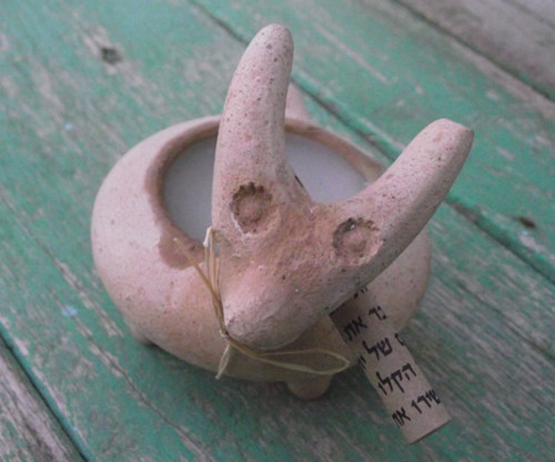 Puede incluir: Una maceta de terracota con forma de animal estilizado con una vela blanca en su interior. La maceta tiene una peque&ntilde;a etiqueta adjunta con texto en hebreo.