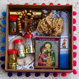 May include: A brown box with a colorful trim contains a variety of religious items, including a rosary, a small icon, a candle, and several vials of holy water, oil, and incense. The box is filled with shredded paper and the text "Святая Земля Иерусалим" is visible on the candle.