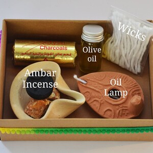 May include: A brown cardboard box with a colorful trim contains a set of incense burning supplies. The box includes a small clay oil lamp, a small bottle of olive oil, a package of wicks, a block of incense, and a pack of charcoals labeled "Charcoals".