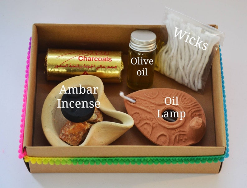 May include: A brown cardboard box with a colorful trim contains a set of incense burning supplies. The box includes a small clay oil lamp, a small bottle of olive oil, a package of wicks, a block of incense, and a pack of charcoals labeled "Charcoals".