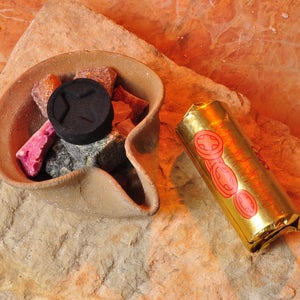 May include: A terracotta incense burner with a round opening filled with small pieces of incense and a black charcoal disc. A gold incense stick with red and gold designs is lying next to the burner.