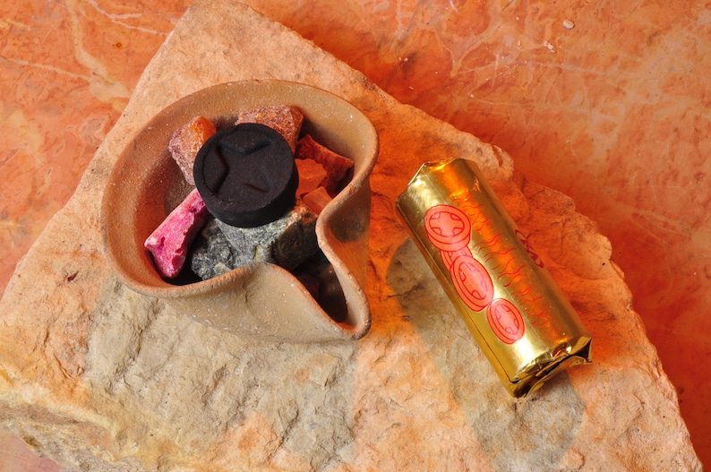 May include: A terracotta incense burner with a round opening filled with small pieces of incense and a black charcoal disc. A gold incense stick with red and gold designs is lying next to the burner.