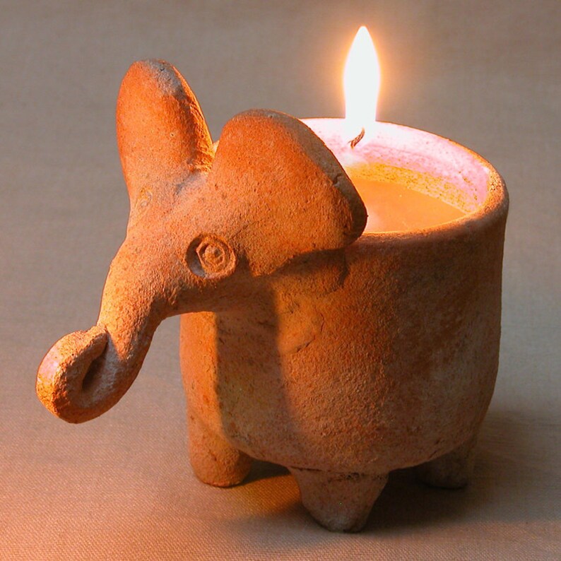 Puede incluir: Un portavelas de terracota con forma de elefante con una vela encendida en su interior. El portavelas tiene una superficie texturizada y un peque&ntilde;o detalle de ojo.