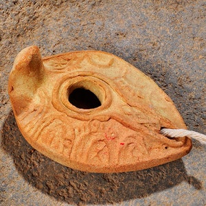 May include: A terracotta oil lamp with a decorative design and a wick. The lamp is sitting on a rough, brown surface.