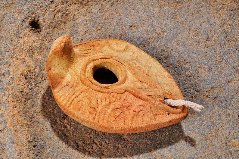May include: A terracotta oil lamp with a decorative design and a wick. The lamp is sitting on a rough, brown surface.