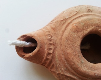 Könnte beinhalten: Eine terracotta-Öllampe mit einem weißen Docht. Die Lampe hat einen runden Körper mit einer kleinen Öffnung für den Docht. Die Lampe ist mit einem einfachen Design verziert.